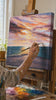 Oslikana zidna slika WEST SLNKA 1-dijelna 70x60 cm. Ručno slikan original na platnu, autentičan interijerni dekor, žena promatra djelo u stilskom svijetlom interijeru. Papilora — Crafted for Home (CFAZH6434E1).